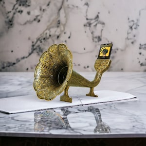 May include: A gold-colored vintage gramophone with intricate details is used as a phone stand. A smartphone is placed in the horn of the gramophone, playing music.