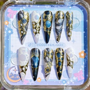 Puede incluir: Un conjunto de diez uñas postizas largas y puntiagudas. Cada uña presenta un diseño único con filigranas doradas, patrones veteados azules y blancos y detalles. Las uñas se exhiben en una caja de plástico transparente con un fondo azul claro y el texto "@ODDream.nails".