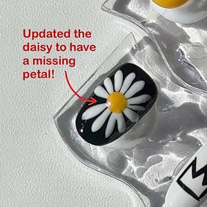 May include: Close-up of nail art designs. One nail features a daisy design with a missing petal, set against a black background. Other nails display a yellow smiley face and a black and white crown. The text "Updated the daisy to have a missing petal!" is also visible.