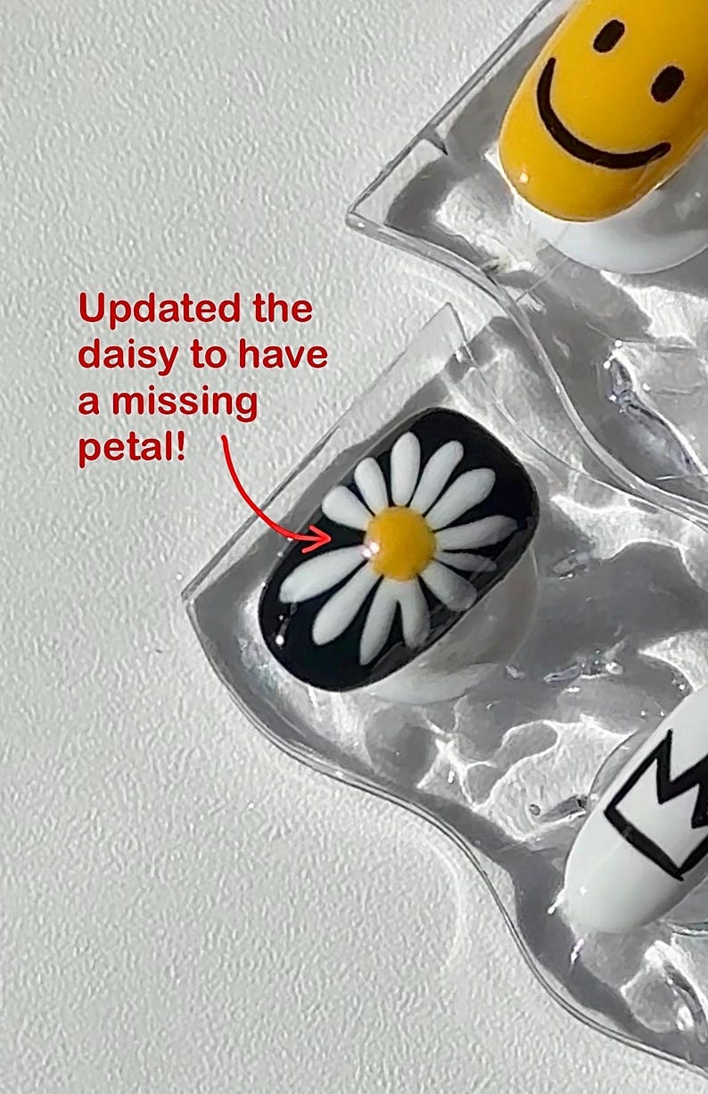May include: Close-up of nail art designs. One nail features a daisy design with a missing petal, set against a black background. Other nails display a yellow smiley face and a black and white crown. The text "Updated the daisy to have a missing petal!" is also visible.