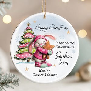 Puede incluir: Un adorno navideño redondo con un conejito de dibujos animados vestido de rosa y sosteniendo una galleta en forma de estrella. El adorno tiene el texto "Happy Christmas" y "To Our Amazing Granddaughter Sophia 2025". También aparece un árbol de Navidad.