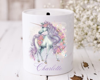 May include: White ceramic piggy bank with a unicorn illustration. The unicorn is white with a purple mane and tail, and is adorned with pink and purple flowers. The piggy bank has a slot on the top for coins and the name "Charlotte" is written on the bottom.
