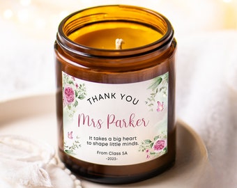 Op de afbeelding: Een bruine glazen kaars met een wit label waarop staat: "Bedankt mevrouw Parker Het kost een groot hart om kleine geesten te vormen. Van klas 5A -2023-." Het label heeft een bloemmotief met roze rozen en vlinders.