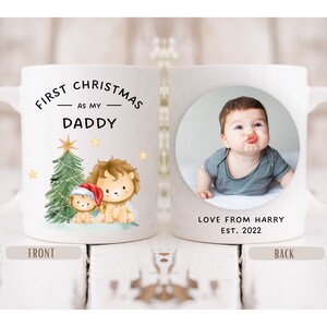 Puede incluir: Taza de cerámica blanca con un diseño navideño con dos leones y un árbol de Navidad. El texto en la taza dice "First Christmas as my Daddy". La parte posterior de la taza tiene una foto de un bebé y el texto "Love from Harry Est. 2022".