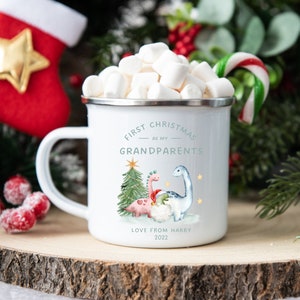 Op de afbeelding: Witte emaille mok met een kerstontwerp met twee dinosaurussen, een kerstboom en de tekst "Eerste Kerstmis als mijn grootouders. Liefs van Harry 2022".