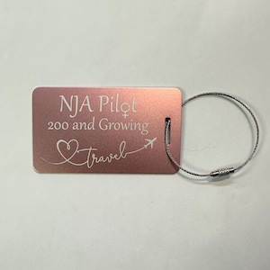 May include: A pink luggage tag with a silver metal loop. The tag has the text "NJA Pilot 200 and Growing" and a heart with a plane and the word "travel" engraved on it.