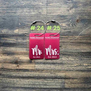 May include: Two red luggage tags with silver rings. The left tag says "Mr. Est 2023" and the right tag says "Mrs. Est 2023". The tags are numbered #24 and #25.
