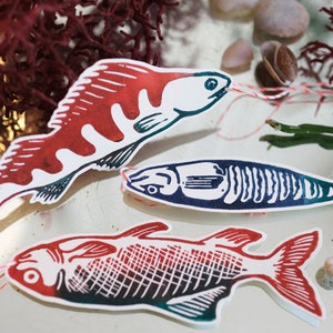 Könnte beinhalten: Drei Papierfische als Dekoration in den Farben Rot, Weiß und Blau. Die Fische sind ausgeschnitten und haben eine Schnur daran befestigt. Die Fische liegen auf einer weißen Oberfläche mit Seegras und Muscheln.