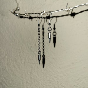 May include: Three silver earrings hanging from a barbed wire fence. The earrings are all different styles, one is a chain with a bullet-shaped charm, one is a bullet-shaped charm with a small ring, and the other is a bullet-shaped charm with a small circle.