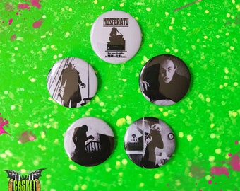 May include: Five black and white button pins featuring scenes from the 1922 German Expressionist horror film "Nosferatu", a silent film adaptation of Bram Stoker's Dracula. The pins feature the title "Nosferatu" and the text "Der neue Grauenfilm - Prana-Film G.m.b.H."