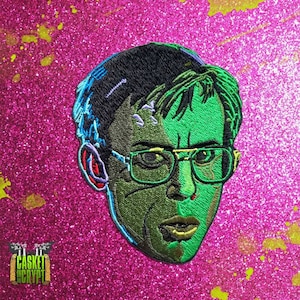 Op de afbeelding: Groen en zwart geborduurd patch van een man met een bril en een serieuze uitdrukking. De patch is op een roze, glinsterende achtergrond.