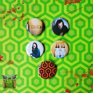 Puede incluir: Conjunto de cinco botones redondos con imágenes de la película The Shining. Los botones presentan una puerta roja con la palabra "REDRUM" escrita en ella, un hombre con una expresión enloquecida, dos chicas con vestidos azules y un patrón repetido de hexágonos rojos y negros.