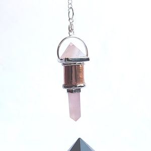 May include: A rose quartz pendulum with a copper and silver base hangs above a clear crystal pyramid with a copper coil inside. The pyramid sits on a wooden base with a flower of life design.