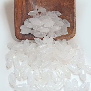 May include: A small, square, dark brown wooden bowl filled with a collection of small, clear, double-terminated quartz crystals. Many of these translucent, faceted crystal points are spilling out of the bowl onto the surface below.