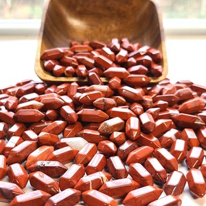 May include: A collection of small, double-terminated red jasper crystal points scattered across a surface, with many pieces spilling out from a wooden bowl. The stones are opaque, reddish-brown in color, and feature a polished, faceted finish.