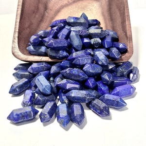 May include: A collection of small, double-terminated lapis lazuli crystals with deep blue coloring and gold-colored flecks, spilling out from a square-shaped wooden bowl with a visible grain pattern and a smooth, polished finish.
