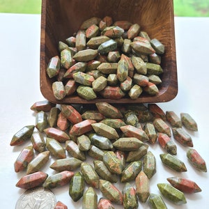 May include: A collection of small, double-terminated unakite crystals with mottled green and pink patterns, contained within a square wooden bowl and scattered on a surface next to a United States quarter coin for scale reference.