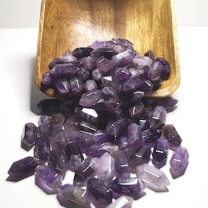May include: A wooden bowl tilted forward, spilling a collection of small, double-terminated amethyst crystals. The crystals vary in shades of deep purple and translucent lavender, featuring pointed ends and faceted surfaces scattered across the surface.