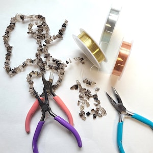 May include: A close-up of jewelry making supplies, including a strand of brown gemstone beads, three spools of wire in silver, gold, and copper, and three pairs of pliers in pink, purple, and blue.