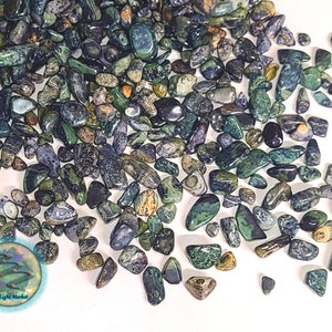 May include: A large pile of green, blue, and black tumbled stones. The stones are various sizes and shapes. The stones are scattered on a white surface. Opal Light Market is printed on a sticker in the lower left corner of the image.