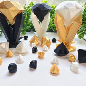 May include: Three 3D printed diamond-shaped objects, each with a different color: gold, black, and white. The diamonds are displayed on a white surface with smaller, scattered diamonds in gold, black, and white.