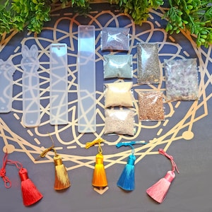 May include: A set of four clear resin bookmark molds, each with a different design. The molds are arranged on a gray background with a mandala pattern. There are also eight small bags of colorful glitter and beads, which are used to fill the molds.