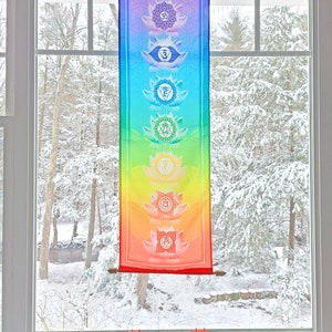 May include: A colorful tapestry with seven chakras, each represented by a different color and symbol, hangs in a window with a snowy view. The tapestry is made of fabric and has a wooden dowel at the top and bottom.