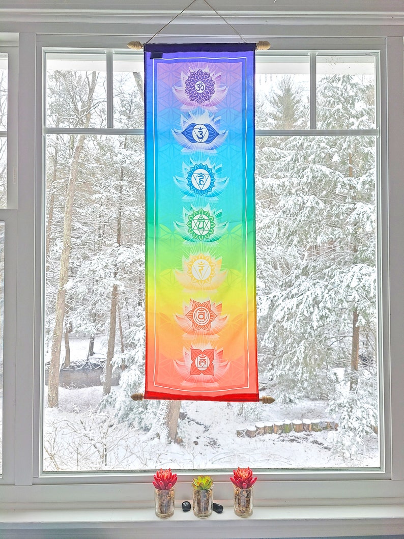 May include: A colorful tapestry with seven chakras, each represented by a different color and symbol, hangs in a window with a snowy view. The tapestry is made of fabric and has a wooden dowel at the top and bottom.