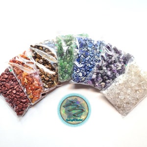 May include: Seven small bags of assorted tumbled gemstones, each bag containing a different color of stone. The bags are arranged in a rainbow pattern, with red, orange, yellow, green, blue, purple, and clear stones. The bags are on a white background. OpalLightMarket is printed on a sticker in the center of the image.