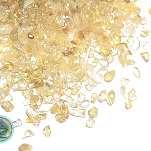 May include: A close-up image of a pile of small, translucent, yellow-gold, tumbled citrine stones. The stones are scattered on a white background.