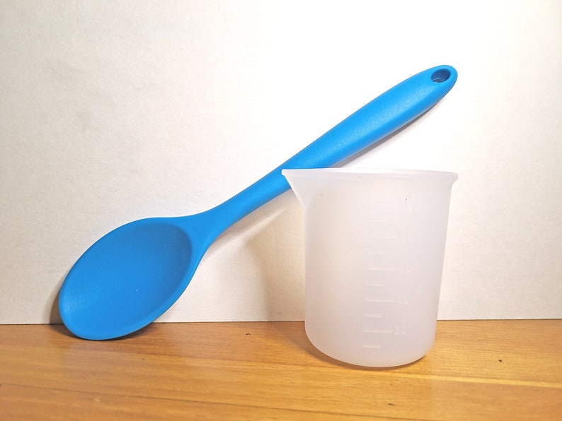 May include: A blue silicone spoon and a white silicone measuring cup with markings on the side. The spoon and cup are on a wooden surface.