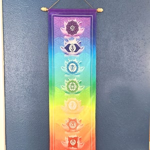 May include: A colorful wall tapestry with seven chakras, each represented by a lotus flower and a symbol. The chakras are arranged vertically from bottom to top, with the root chakra at the bottom and the crown chakra at the top. The tapestry is made of fabric and has a wooden dowel at the top and bottom.