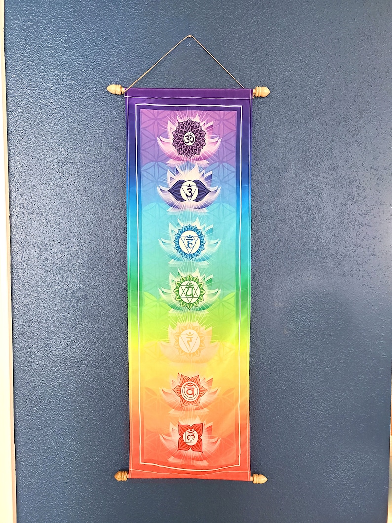 May include: A colorful wall tapestry with seven chakras, each represented by a lotus flower and a symbol. The chakras are arranged vertically from bottom to top, with the root chakra at the bottom and the crown chakra at the top. The tapestry is made of fabric and has a wooden dowel at the top and bottom.