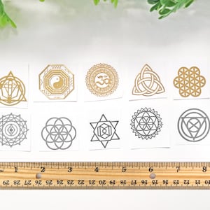 May include: Ten gold and silver temporary tattoos with geometric designs, including a flower of life, a yin yang symbol, and a Celtic knot. The tattoos are arranged in two rows of five on a white background.