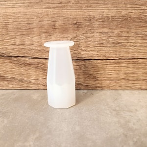 May include: A translucent white bottle stopper with a hexagonal base and a flat, circular top. The stopper has a tapered, faceted body. The background is a wooden surface.