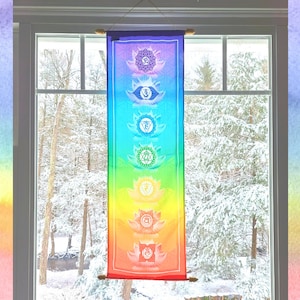 May include: A colorful tapestry with seven chakras, each represented by a different color and symbol, hangs in a window with a snowy view.