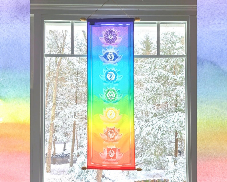 May include: A colorful tapestry with seven chakras, each represented by a different color and symbol, hangs in a window with a snowy view.