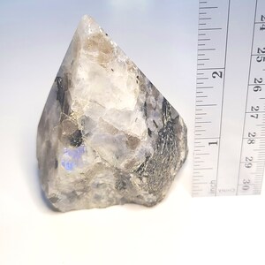 May include: A large, clear, iridescent moonstone crystal point with black inclusions. The crystal is approximately 2 inches tall and is sitting on a white surface next to a ruler.