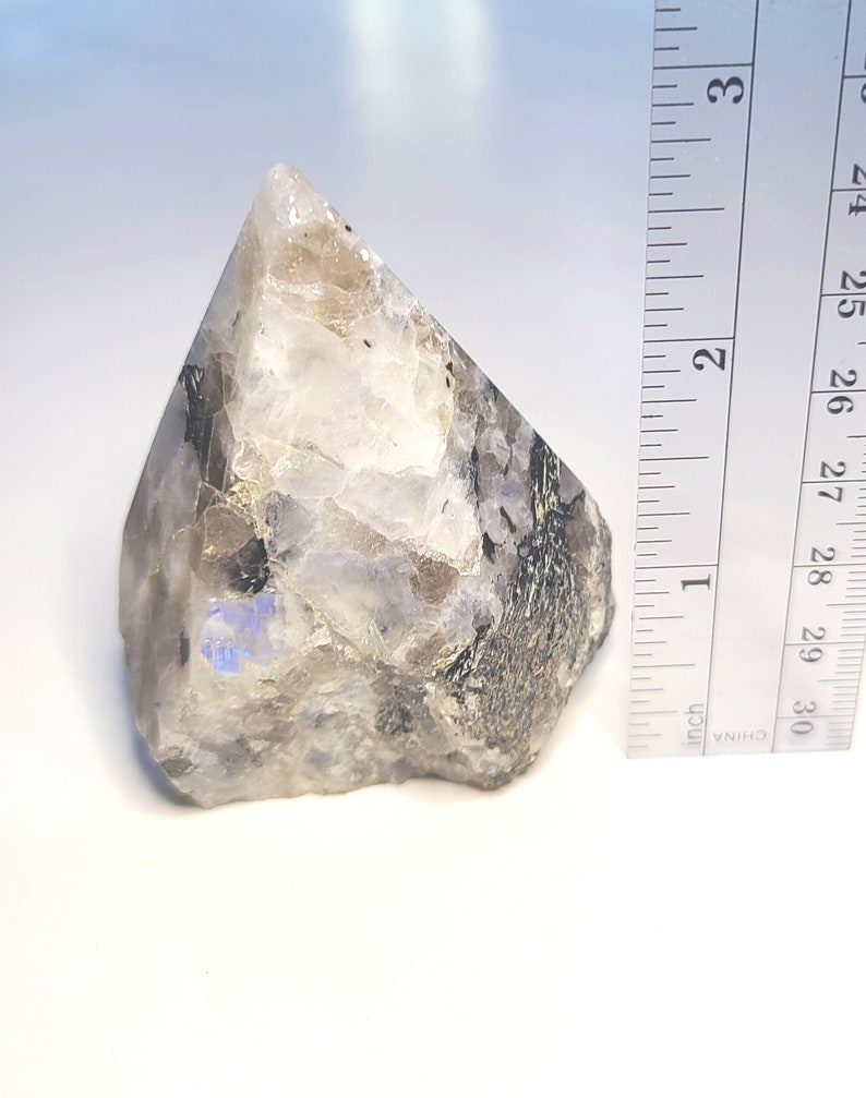 May include: A large, clear, iridescent moonstone crystal point with black inclusions. The crystal is approximately 2 inches tall and is sitting on a white surface next to a ruler.