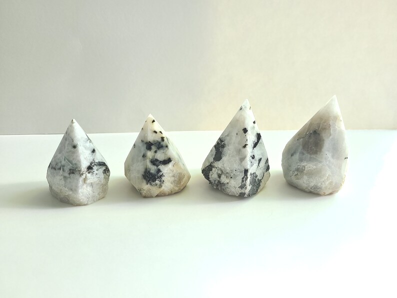May include: Four white and black gemstone points, also known as rainbow moonstone points, arranged in a row on a white surface.