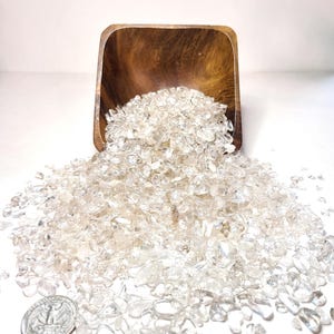 May include: A wooden bowl overflowing with clear quartz crystals, spilling onto a white surface. A US quarter is included for scale.