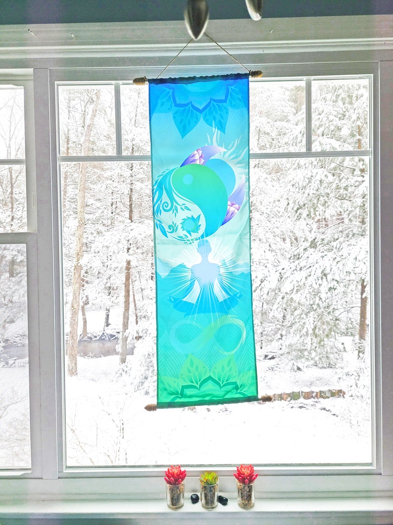 May include: A blue and green tapestry with a yin yang symbol and a meditating figure. The tapestry is hanging in a window with a snowy view outside.