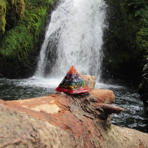 May include: A colorful crystal pyramid sits on a log in front of a waterfall. The pyramid is made of various crystals, including amethyst, clear quartz, and citrine. The waterfall is surrounded by lush green foliage.