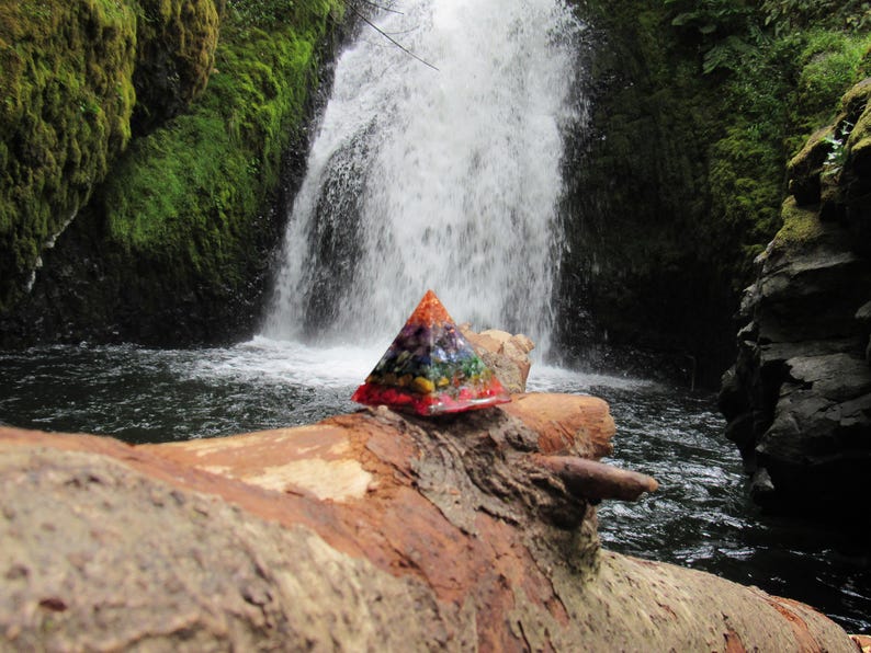 May include: A colorful crystal pyramid sits on a log in front of a waterfall. The pyramid is made of various crystals, including amethyst, clear quartz, and citrine. The waterfall is surrounded by lush green foliage.