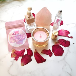 May include: An assortment of self-care products, including a pink herbal tea packet, a rose quartz crystal, a glass candle holder with a lit pink candle, a roller bottle, and a small gold-topped bottle. Red rose petals are scattered around. The tea packet has text.