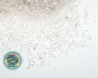 May include: A close-up of a pile of small, clear quartz crystals scattered on a white background. The crystals are various shapes and sizes, with some having a slightly iridescent sheen.