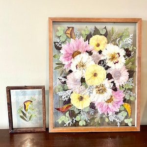 May include: Two rectangular wooden frames containing pressed flowers. The larger frame features a dense arrangement of pink, yellow, and white flowers with green and silver foliage. The smaller frame holds a single calla lily with green leaves.