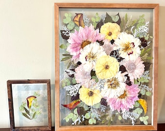 May include: Two rectangular floating frames with wooden borders containing pressed flowers. The larger frame features a dense arrangement of pink, yellow, and white flowers with green foliage. The smaller frame holds a single yellow calla lily with leaves.