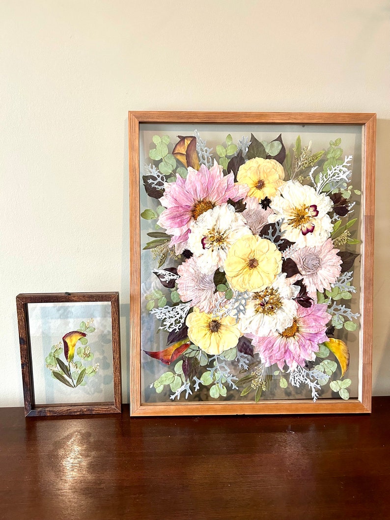 May include: Two rectangular wooden frames containing pressed flowers. The larger frame features a dense arrangement of pink, yellow, and white flowers with green and silver foliage. The smaller frame holds a single calla lily with green leaves.