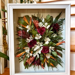 May include: A framed pressed flower arrangement with a variety of dried flowers and leaves, including red roses, white flowers, orange and green leaves, and brown ginger flowers.